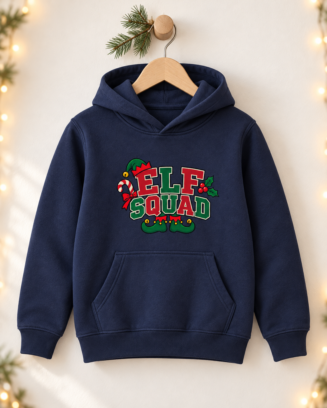 A navy blue kids' pullover hoodie hanging on a wooden hanger against a neutral wall with pine branches and warm fairy lights. The hoodie features a festive "ELF SQUAD" graphic on the chest in red, green, and white lettering, accented with a candy cane, holly leaves, and elf shoes.