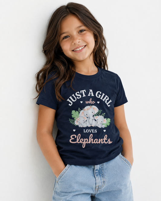 A young girl with long brown hair smiling and leaning against a white wall, wearing a navy blue graphic t-shirt with a cartoon elephant design and the text 'JUST A GIRL who LOVES Elephants', paired with light blue jeans.