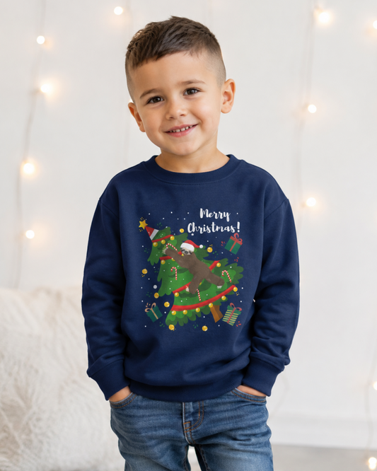 Young boy wearing a navy kids Christmas sweatshirt featuring a sloth in a Santa hat stretched across a decorated Christmas tree with candy canes, gifts and Merry Christmas text.