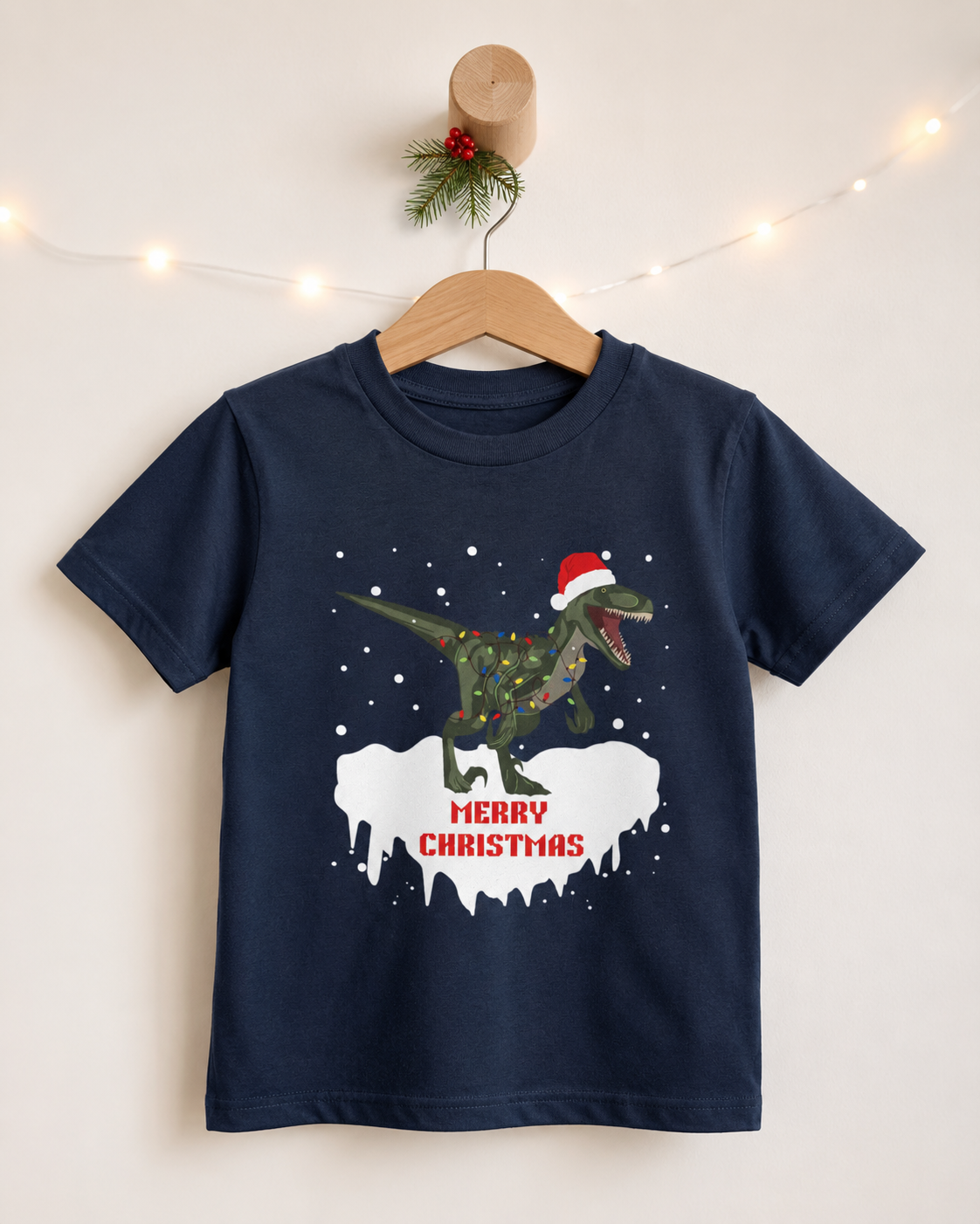 Navy kids Christmas T-shirt on a wooden hanger featuring a roaring T-Rex in a Santa hat wrapped in colourful Christmas lights, with falling snow and Merry Christmas text.