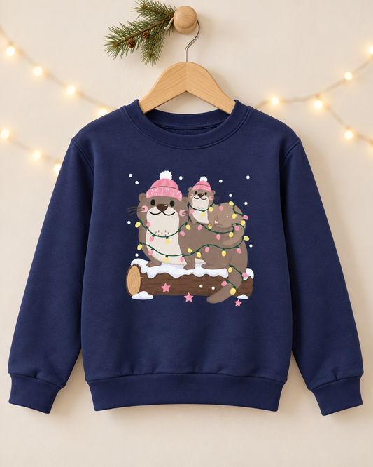A kids navy blue crewneck sweatshirt hanging on a wooden hanger against a warm beige background with soft string lights and a pine branch. The sweatshirt features a cartoon graphic of two otters wearing pink winter hats and wrapping in holiday lights.