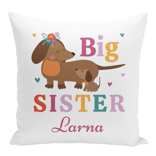 A white square cushion features a cartoon-style illustration of a brown dachshund with a flower crown, accompanied by a smaller dachshund. The text "Big SISTER" is displayed above the dogs in colourful letters, and the name "Larna" is written below in a flowing pink script.