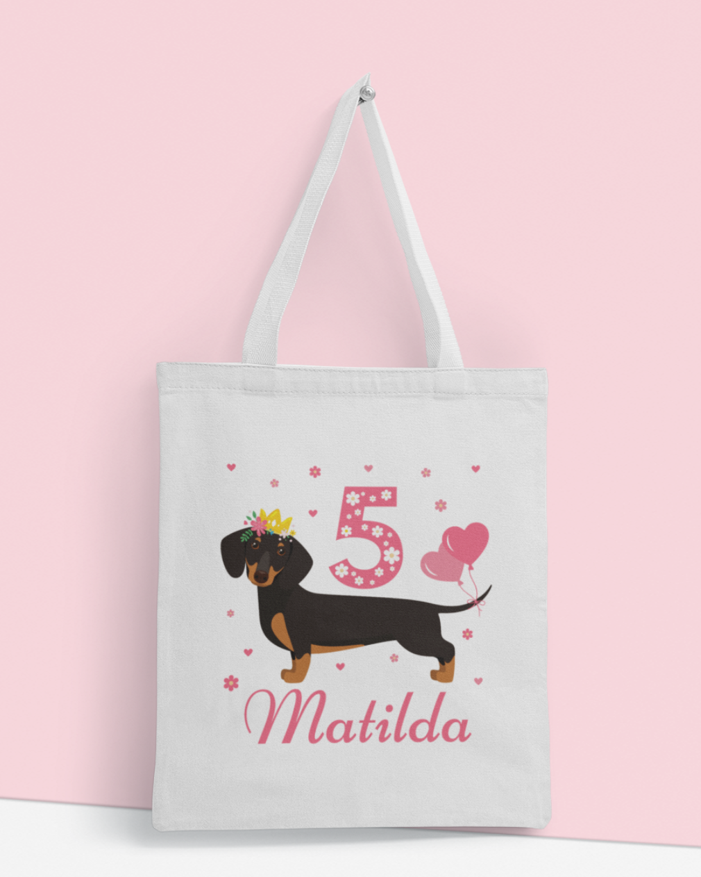 Personalised white birthday tote against a pink wall, printed with a black and tan dachshund in a flowered crown, pink number 5 and the name Matilda.