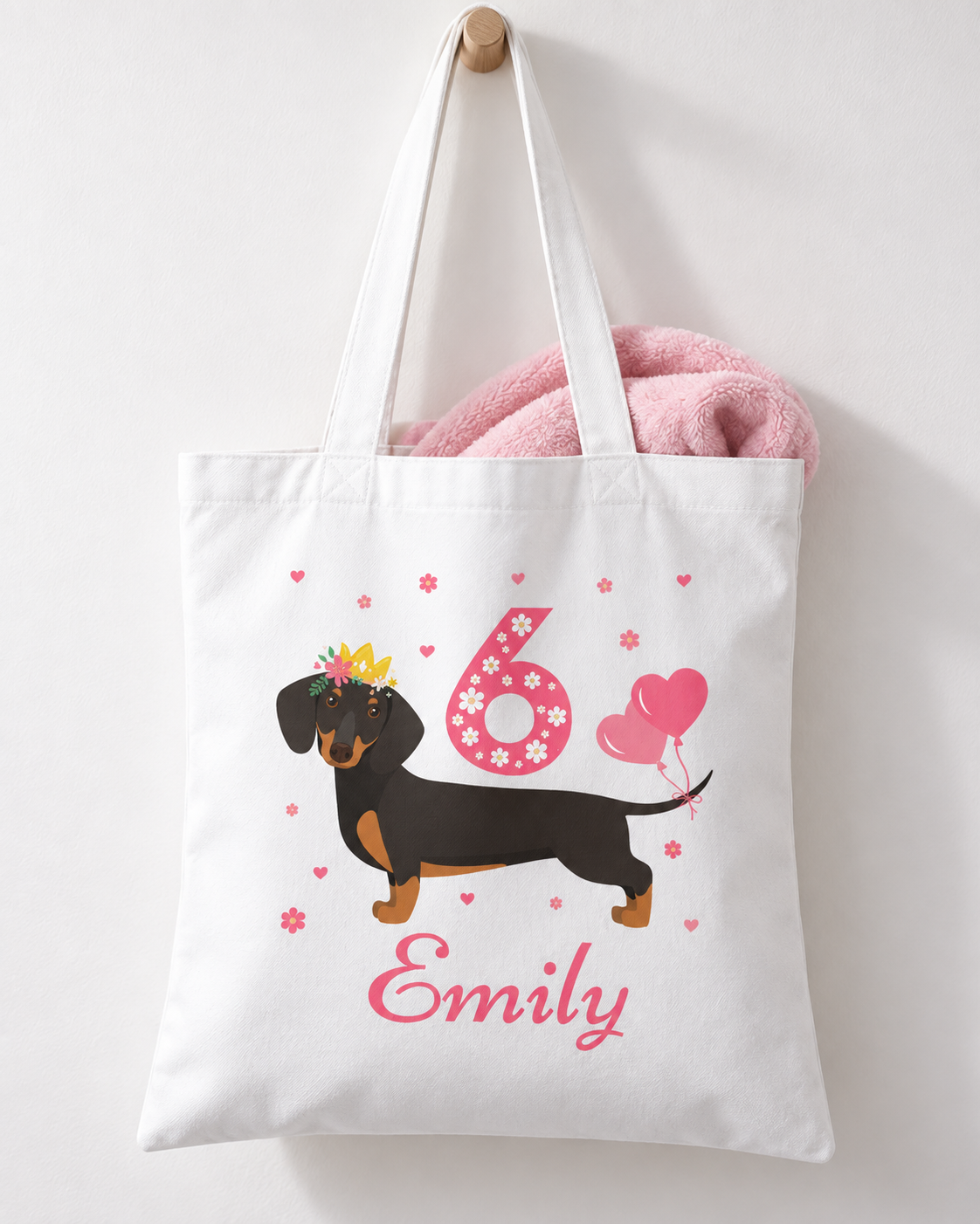A kids white cotton tote bag with long handles, featuring a printed design of a dachshund wearing a yellow crown, a pink number 6 with flowers, pink hearts, and the name 'Emily' in pink script. A pink plush toy is tucked inside the bag, which hangs from a wooden peg on a white wall.
