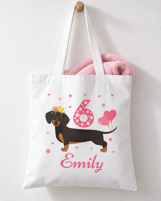 A kids white cotton tote bag with long handles, featuring a printed design of a dachshund wearing a yellow crown, a pink number 6 with flowers, pink hearts, and the name 'Emily' in pink script. A pink plush toy is tucked inside the bag, which hangs from a wooden peg on a white wall.