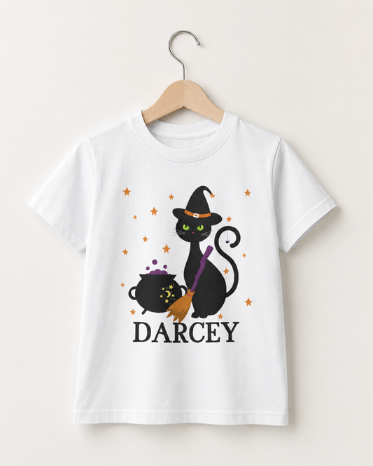 Personalised kids white Halloween T-shirt with black witch cat, bubbling cauldron, broom and the name Darcey, displayed on a wooden hanger.