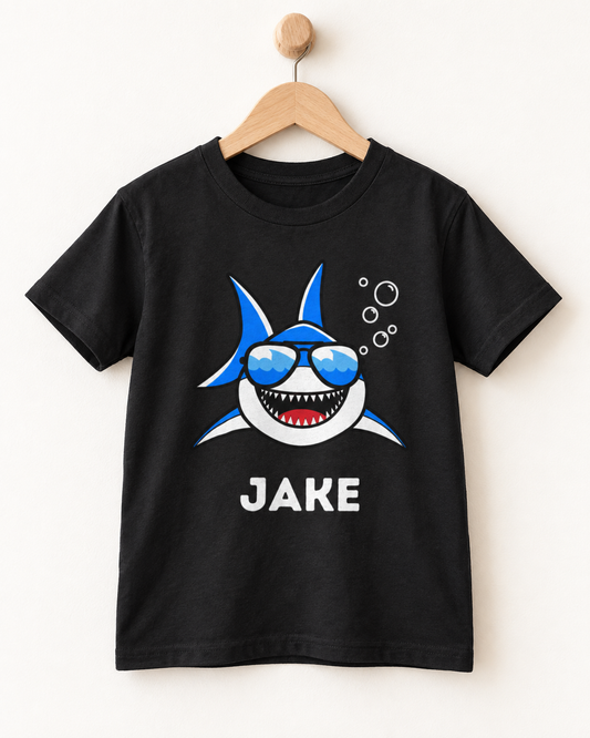 Personalised kids black T-shirt featuring a smiling shark in sunglasses and name, displayed on a wooden hanger and wall hook.