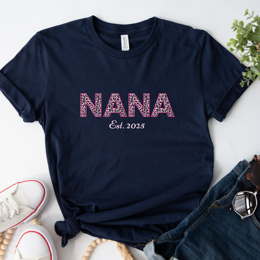 Navy blue t-shirt with 'NANA Est. 2025' text on a white surface with accessories.