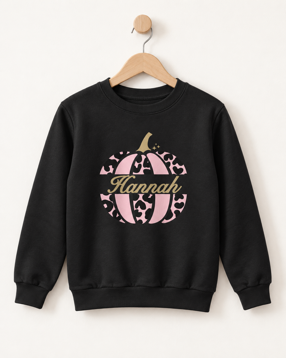A girls black crewneck sweatshirt hanging on a wooden hanger against a plain off-white wall. The sweatshirt features a graphic of a pink leopard-print and striped pumpkin with the name 'Hannah' written in gold script across the center.