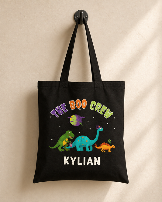 Black personalised cotton tote bag hanging from a wall hook, featuring colourful Halloween dinosaurs, “The Boo Crew” and the name Kylian.