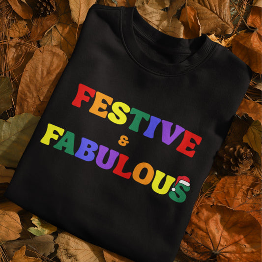 Folded black sweatshirt with Pride rainbow text reading "Festive & Fabulous" and a Santa hat on the letter "S", displayed on autumn leaves and pinecones.