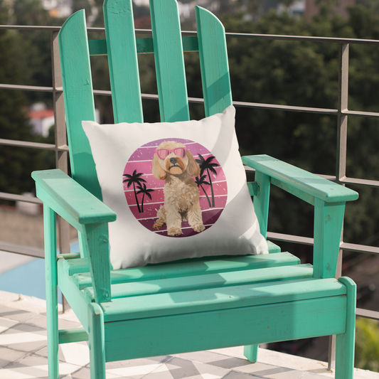A white square cushion with a retro-style print of a cockapoo wearing pink sunglasses, set against a pink and purple striped circle with black palm trees. The cushion is placed on a teal wooden chair on a tiled surface.