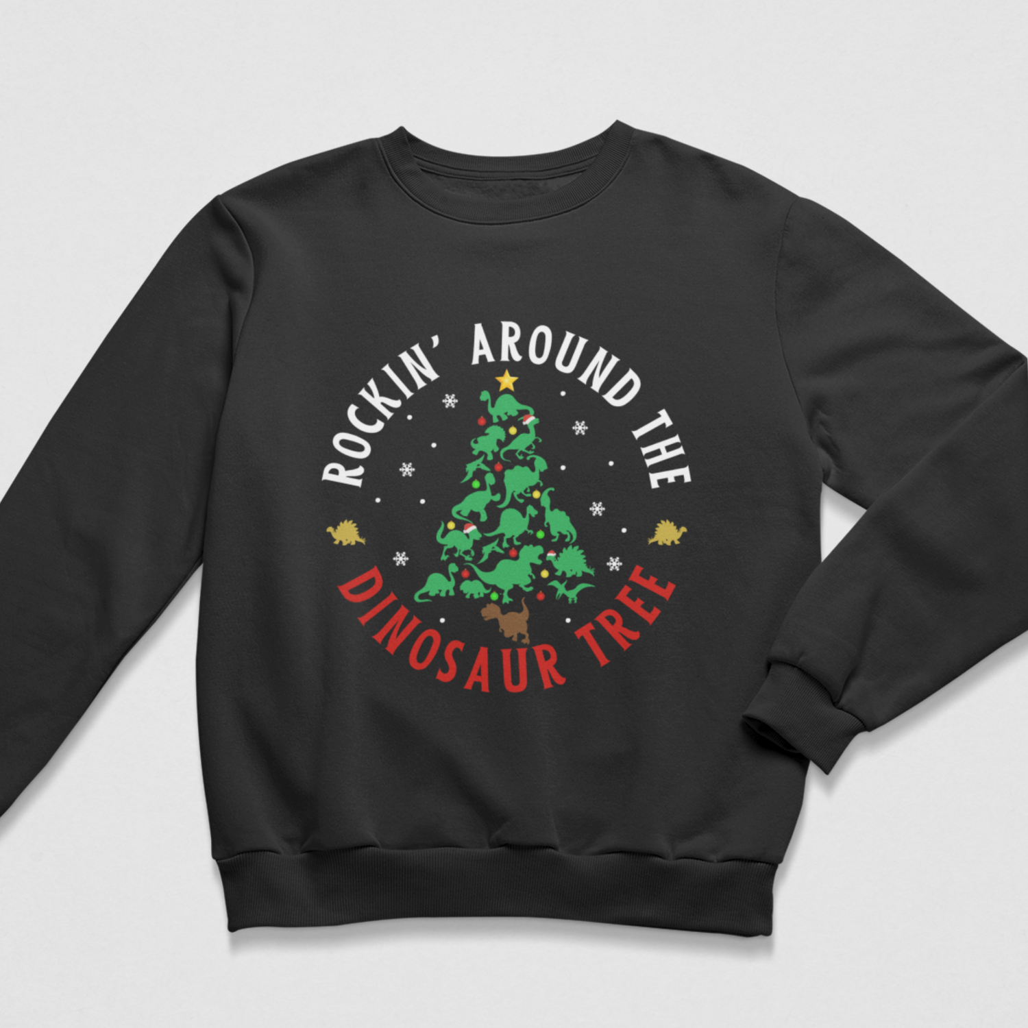 A kids black crewneck sweatshirt featuring a festive graphic print of a Christmas tree surrounded by the text 'ROCKIN' AROUND THE DINOSAUR TREE' in white and red, with small dinosaur silhouettes and snowflakes.