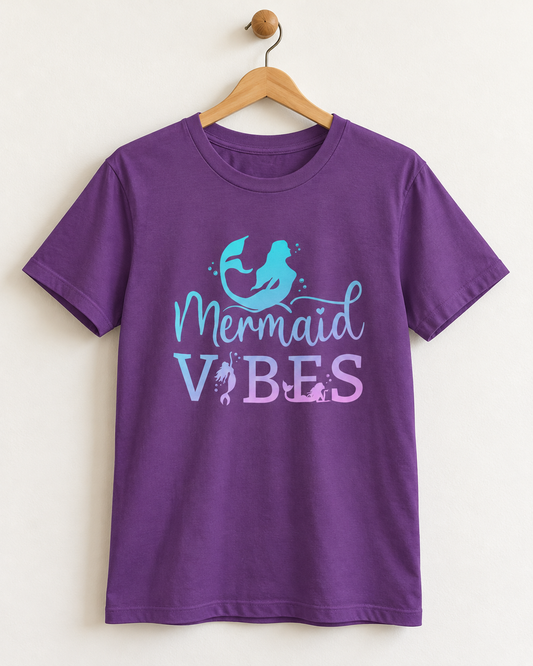 A purple short-sleeve crewneck t-shirt hanging on a wooden hanger against a light beige wall. The shirt features a graphic print on the front with a light blue mermaid tail and mermaid silhouette above the text 'Mermaid VIBES' in light blue and lavender.