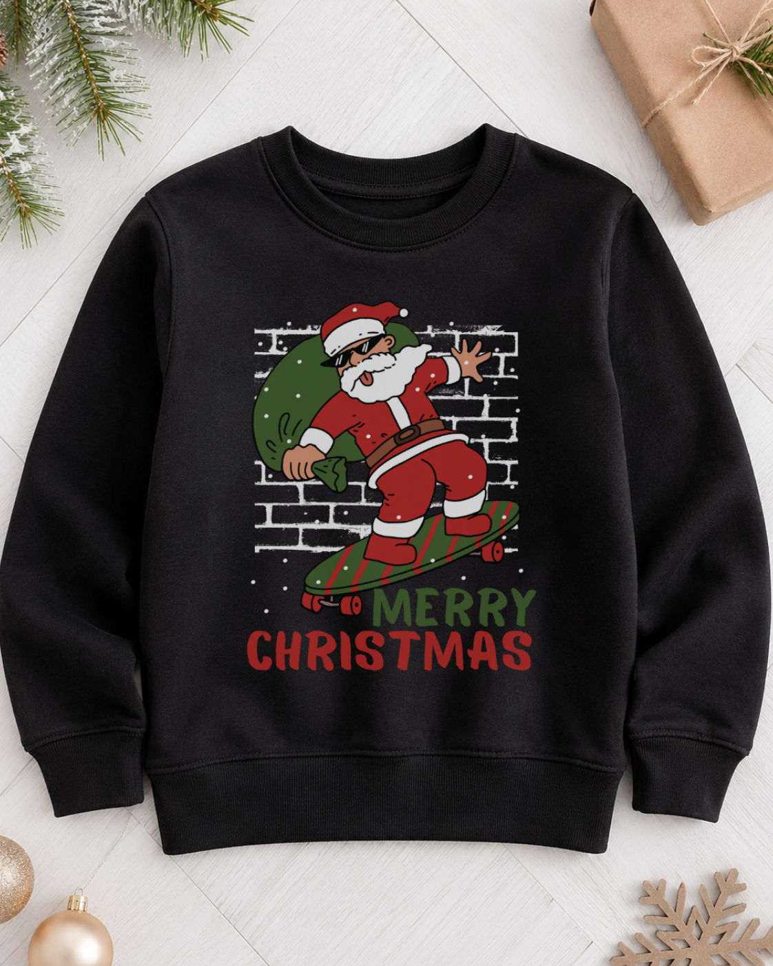 A kids black crewneck sweatshirt featuring a festive graphic of a dancing Santa Claus on a skateboard in front of a brick wall, with the text 'IT'S GO MERRY CHRISTMAS' printed below. The sweatshirt is displayed in a flat-lay setting with holiday decorations including pine branches, a wrapped gift, ornaments, and a wooden snowflake on a white parquet floor.