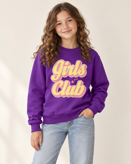 A young girl with long wavy brown hair wearing a purple crewneck sweatshirt with 'Girls Club' printed in pink and yellow bubble letters, paired with light blue jeans in a studio setting.
