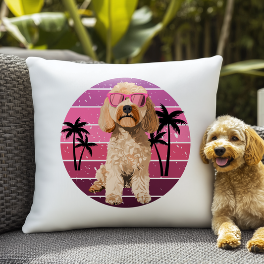 Cockapoo-themed cushion on an outdoor wicker chair, featuring a printed illustration of a cockapoo wearing pink sunglasses with a pink sunset and palm tree background. A real cockapoo sits beside the cushion, summery dog-lover scene.