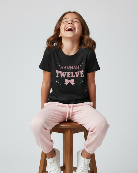 12 year old girl wearing black Hannah Twelve birthday t-shirt with pink bow and sparkles, seated on stool in studio with pink joggers and white trainers