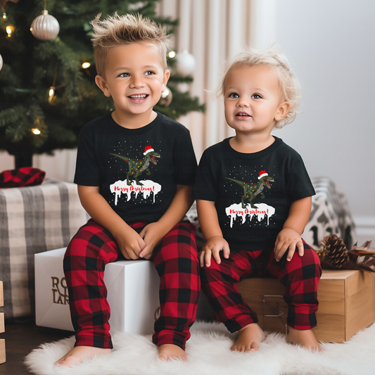 Two children wearing black crew neck T-shirts with a festive T-rex design and red and black checkered pants, sitting in front of a Christmas tree.