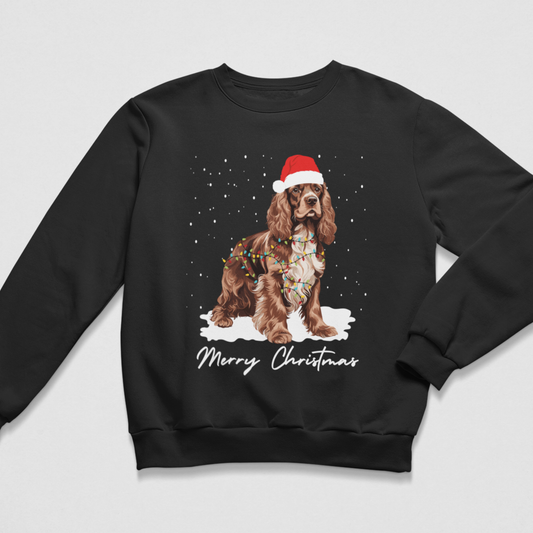 A black crewneck sweatshirt featuring a graphic of a brown and white Cocker Spaniel wearing a red Santa hat and fairy lights, standing in the snow with 'Merry Christmas' written below, displayed on a light grey background.
