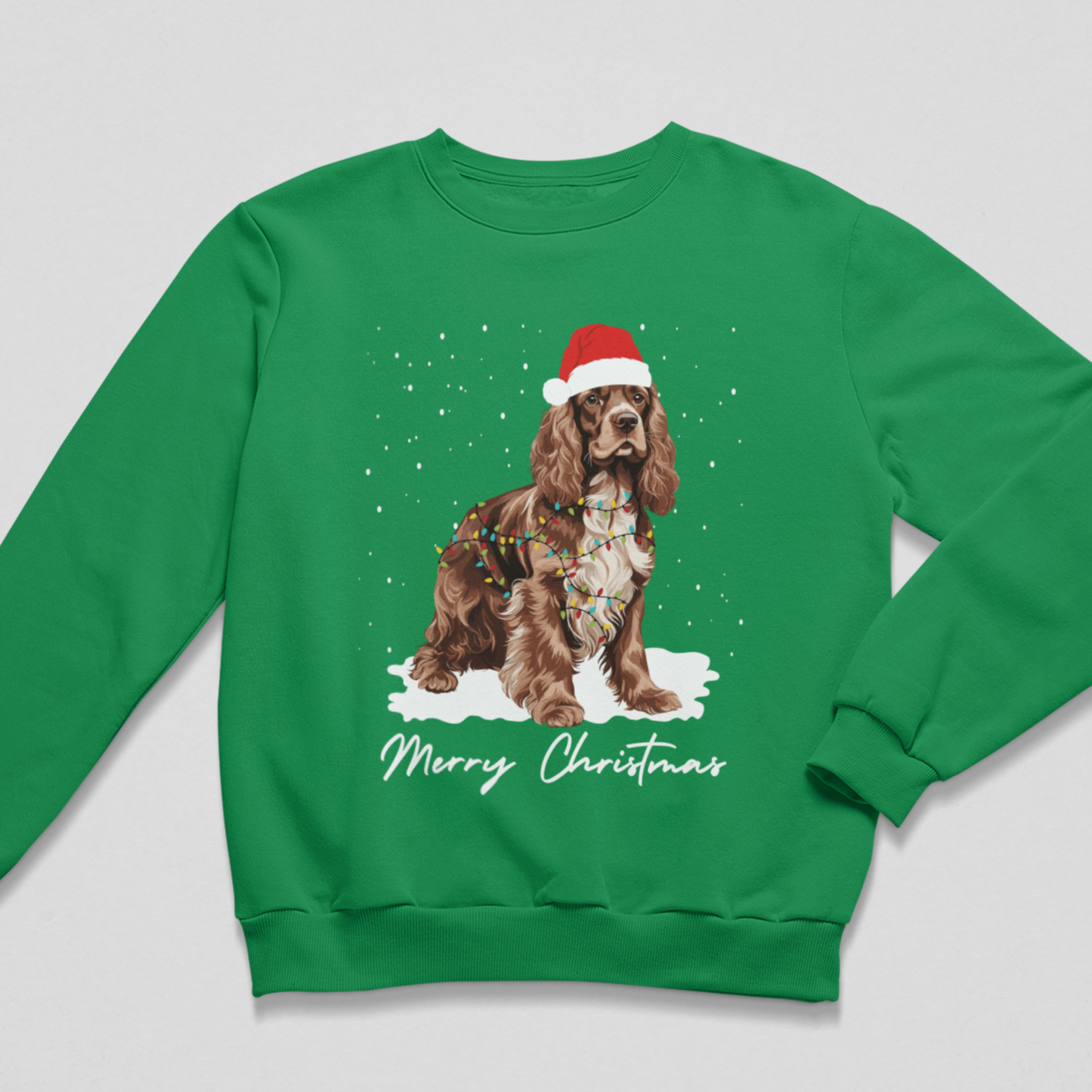 Adults green crewneck sweatshirt featuring a graphic of a brown and white Cocker Spaniel wearing a red Santa hat and Christmas lights, with 'Merry Christmas' written below in white script, shown on a light grey background.
