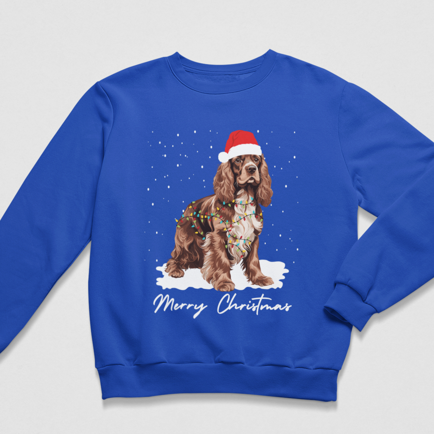 Adults royal blue crewneck sweatshirt featuring a graphic of a brown Cocker Spaniel wearing a red Santa hat and holiday lights, sitting in the snow with 'Merry Christmas' written below, displayed on a light grey background.