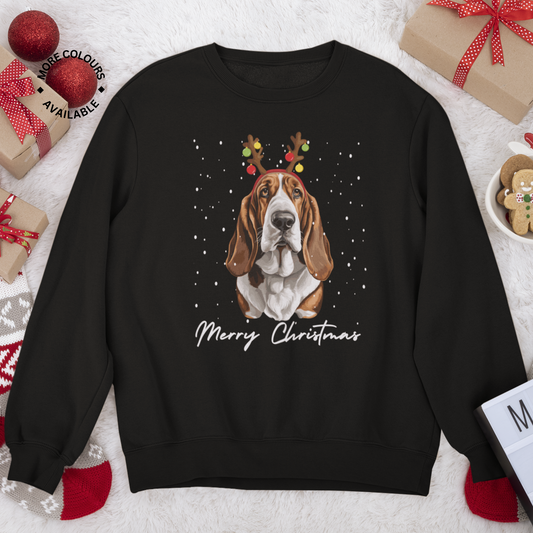Flat lay of black Christmas sweatshirt featuring a festive Basset Hound wearing reindeer antlers and surrounded by falling snow, styled with holiday décor
