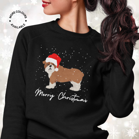 Woman wearing a black Christmas sweatshirt with an English Bulldog in a Santa hat, snow falling, and “Merry Christmas” text below