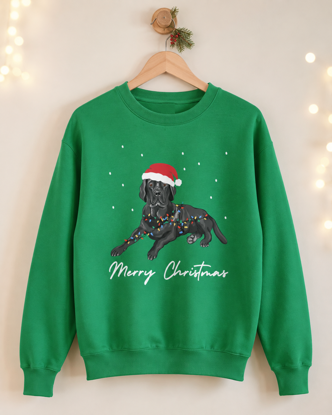 Adults green crewneck sweatshirt hanging on a wooden hanger against a warm beige wall with soft string lights. The sweatshirt features a graphic of a black Labrador wearing a red Santa hat and Christmas lights, with 'Merry Christmas' written in white script below.