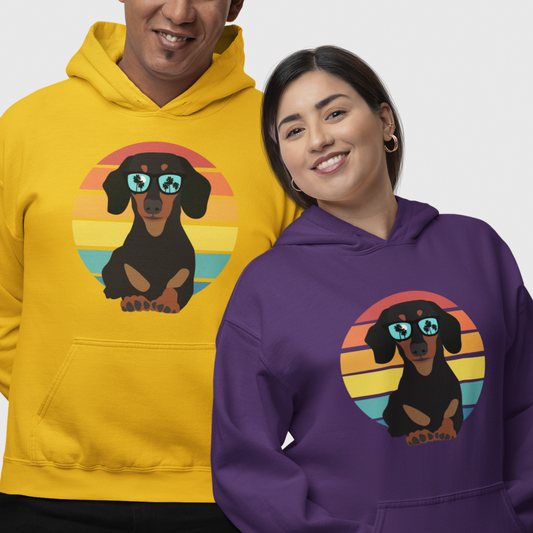 Two smiling adults wear hoodies with a graphic of a dachshund wearing sunglasses against a retro striped circle. The hoodie on the left is yellow, and the one on the right is purple.