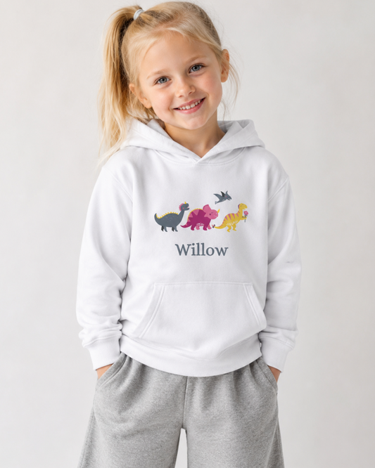 Smiling blonde girl wearing a white personalised dinosaur hoodie with a colourful dinosaur line-up and the name Willow, styled with soft grey wide-leg joggers.