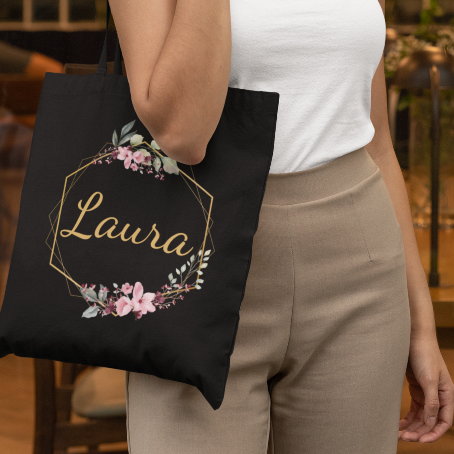 A woman holding a black tote bag with floral gold hexagon frame and personalised name “Laura” in gold script.