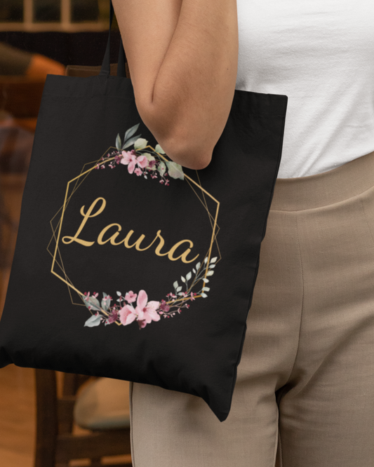 A woman holding a black tote bag with floral gold hexagon frame and personalised name “Laura” in gold script.