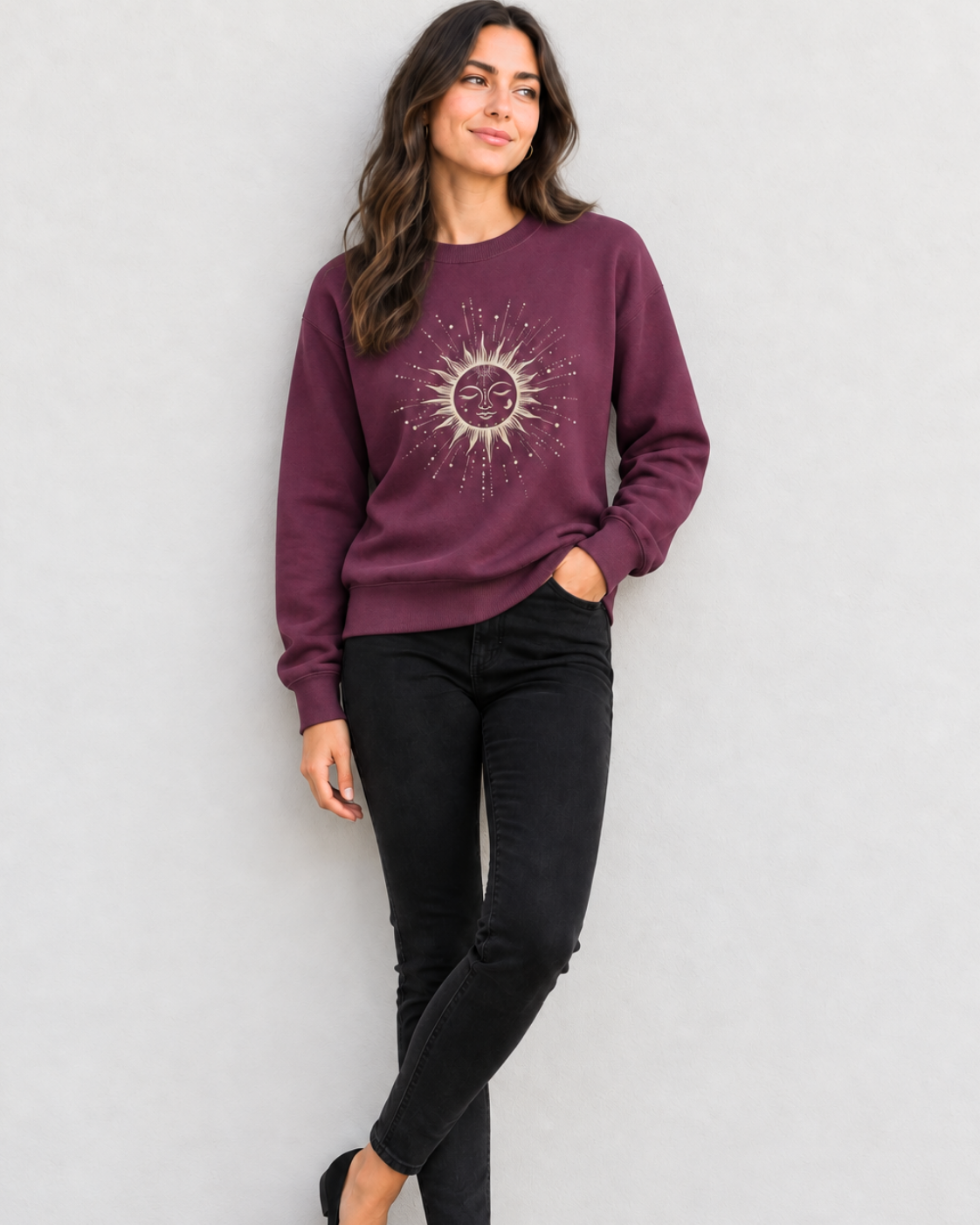Woman wearing a plum coloured crew neck sweatshirt featuring a celestial sun face design paired with black jeans.