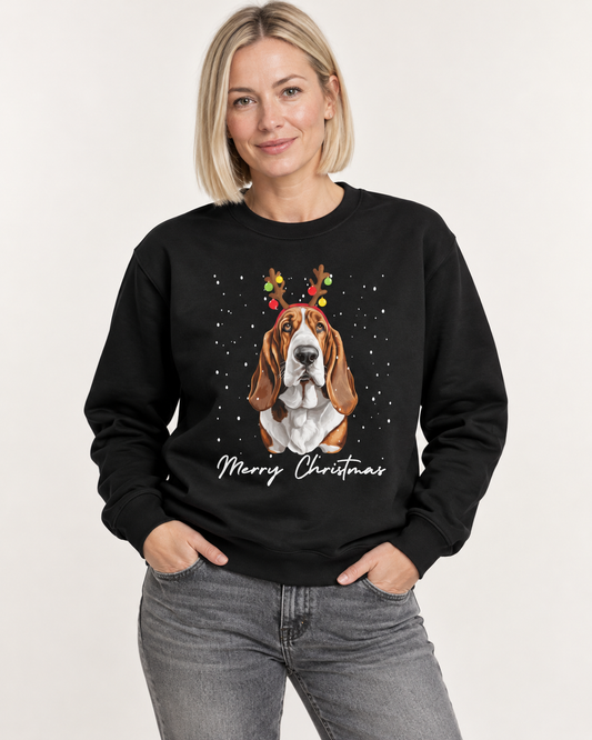 A woman wearing a black crewneck sweatshirt with a graphic of a Basset Hound wearing festive reindeer antlers and the words 'Merry Christmas' printed below, paired with gray jeans against a plain light background.