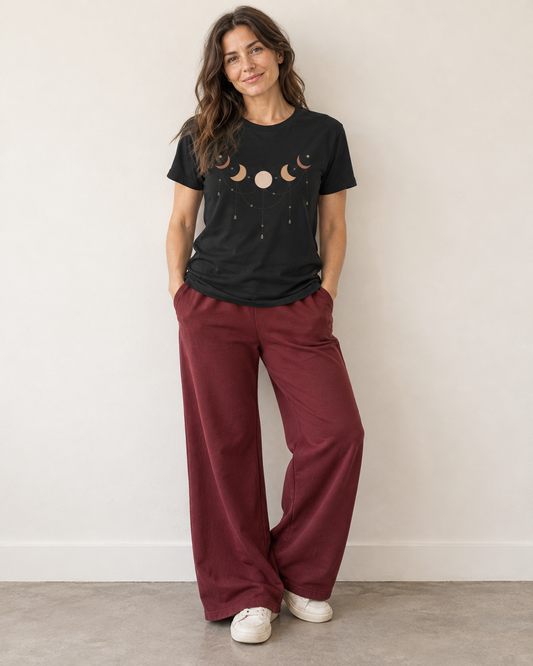 A woman stands against a plain off-white wall wearing a black graphic t-shirt with a moon phase design and wide-leg burgundy joggers.
