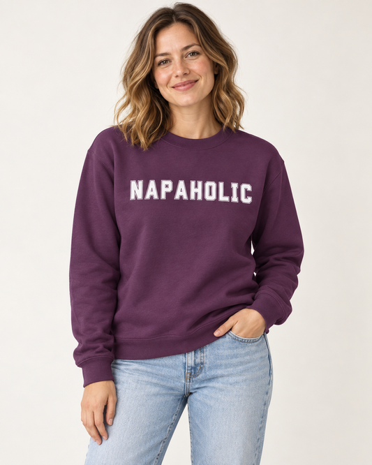A woman wearing a purple crewneck sweatshirt with 'NAPAHOLIC' printed in white across the chest, paired with light blue jeans in a studio setting.