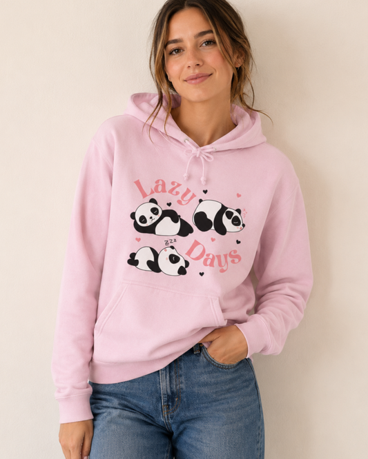 A woman wearing a light pink pullover hoodie with a graphic of three sleeping pandas and the words 'Lazy Panda Days' in pink, paired with blue jeans, standing against a plain beige wall.