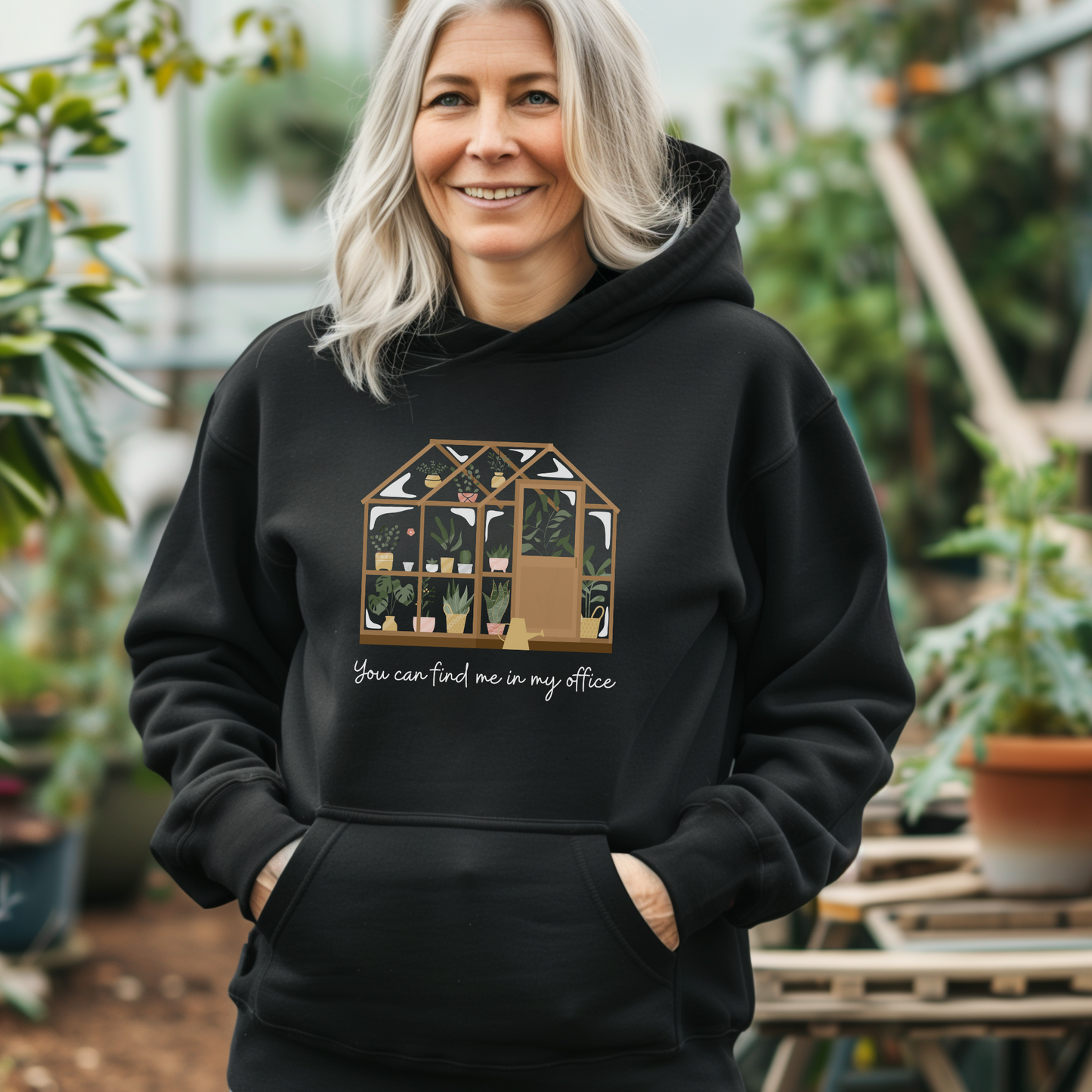 Woman standing in a greenhouse wearing a black hoodie with the printed design of a greenhouse and words saying "You can find me in my office"