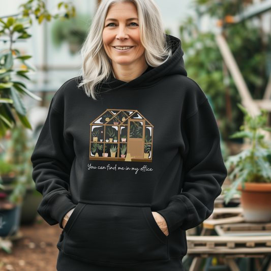 Smiling woman in "You can find me in my office" greenhouse and plants hoodie stands large green house amongst pots and plants.