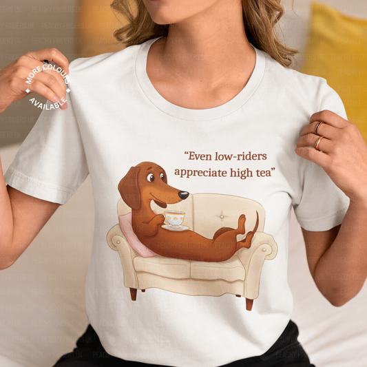 A woman models a white t-shirt featuring a cartoon dachshund lounging on a cream sofa, holding a teacup. The text above reads "Even low-riders appreciate high tea".