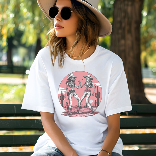 A woman sitting on a bench wearing a white cotton oversized t-shirt with a printed design of cowgirl skeletons dancing in western style with mountains and cacti.