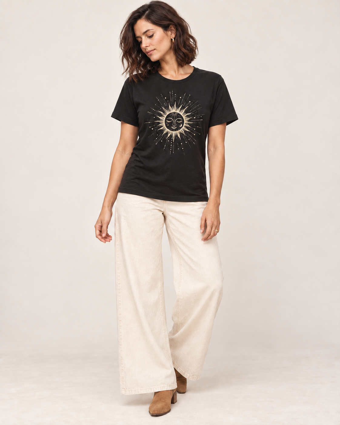 Woman wearing a black celestial sun and moon T-shirt with cream wide-leg trousers and tan ankle boots in a bright studio.