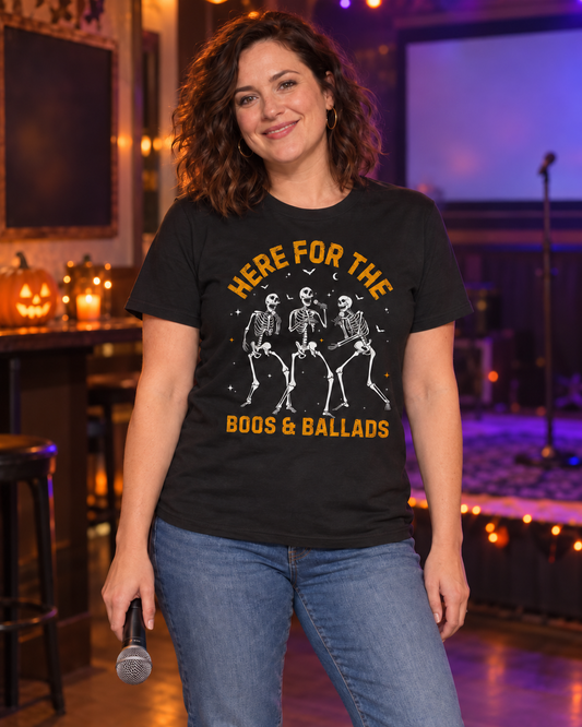Woman at a karaoke night wearing a black Halloween T-shirt featuring three singing skeletons and the words “Here for the Boos & Ballads.”