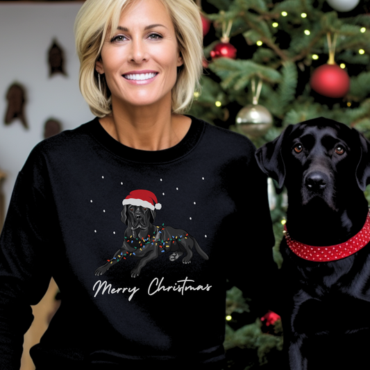 A woman wearing a black festive sweatshirt with an image of a Labrador wearing a Santa hat and wrapped in festive lights, standing next to a black Labrador. The sweatshirt has the text 'Merry Christmas'.