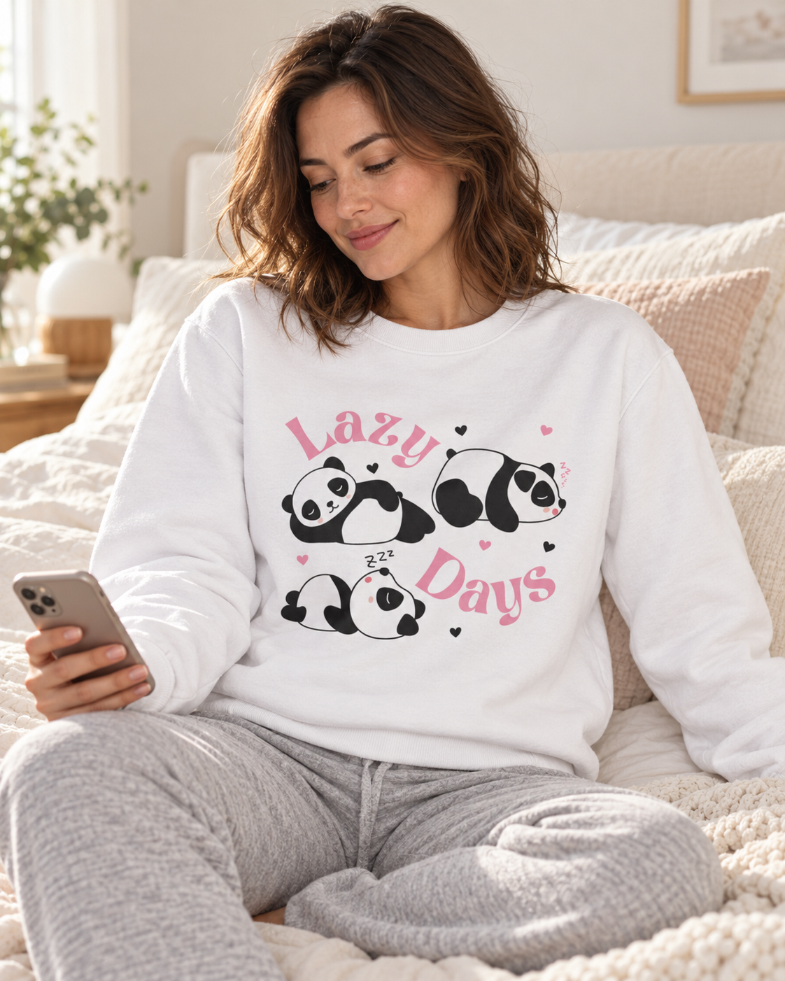 A woman sitting on a bed wearing a white crewneck sweatshirt with a graphic of three sleeping pandas and the words 'Lazy Days' in pink, paired with grry sweatpants.
