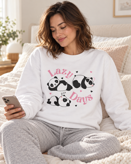 A woman sitting on a bed wearing a white crewneck sweatshirt with a graphic of three sleeping pandas and the words 'Lazy Days' in pink, paired with grry sweatpants.