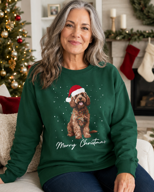 A woman wearing a dark green Christmas sweatshirt featuring a brown Cockapoo in a Santa hat surrounded by falling snow, with “Merry Christmas” script, seated on a sofa in a bright, festively decorated living room.