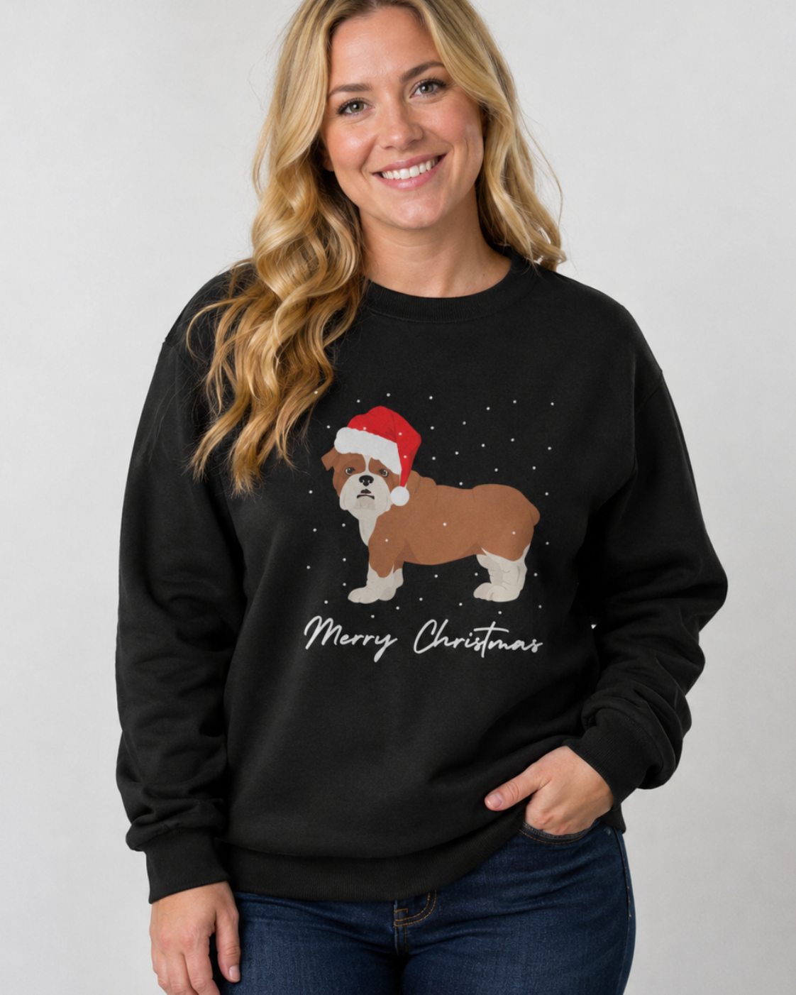 A smiling woman with blonde hair models a black crewneck sweatshirt featuring a cartoon British Bulldog wearing a red Santa hat and falling snow, with 'Merry Christmas' written below in white script.