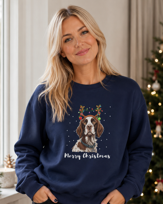 A woman wearing a navy blue crewneck sweatshirt with a Christmas graphic of a Springer Spaniel wearing a reindeer antler crown and the text 'Merry Christmas' underneath, standing in a cozy room with a decorated Christmas tree in the background.